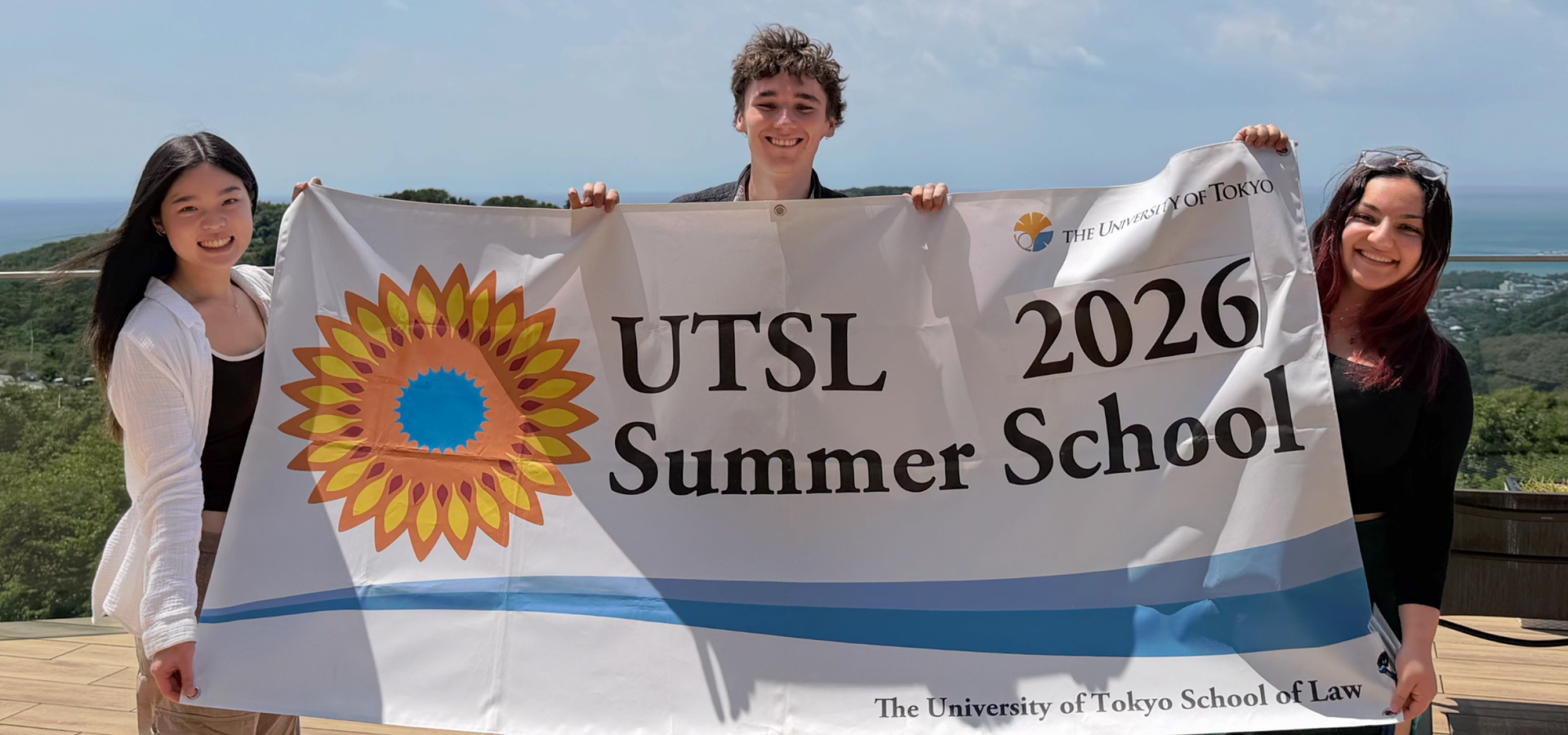 ANU Law students Bita Mahani, William Maxwell and Chloe Law smile while holding a University of Tokyo Law School Summer School 2026 banner, with the Japanese coastline in the background.